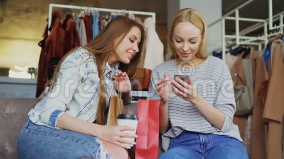 快乐的年轻女性聊天和使用现代智能手机同时坐在漂亮的服装精品店他们微笑着视频的预览图