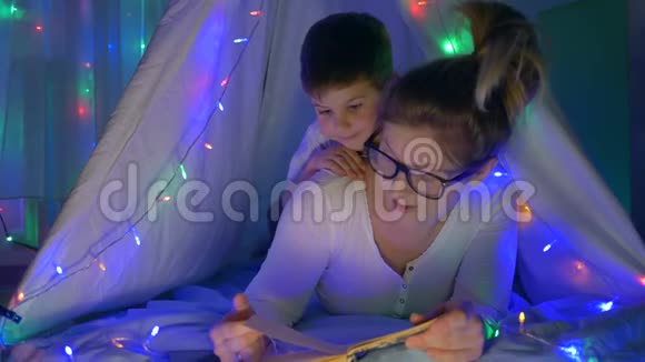 母亲与孩子的关系妈妈戴着眼镜在睡觉前给幼儿看书躺在帐篷里家里灯火辉煌视频的预览图
