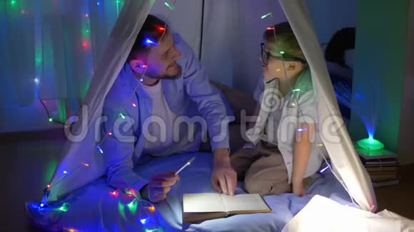 学前教育年轻的父亲拿着手电筒给男孩看童话故事戴上眼镜坐在有花环的帐篷里视频的预览图