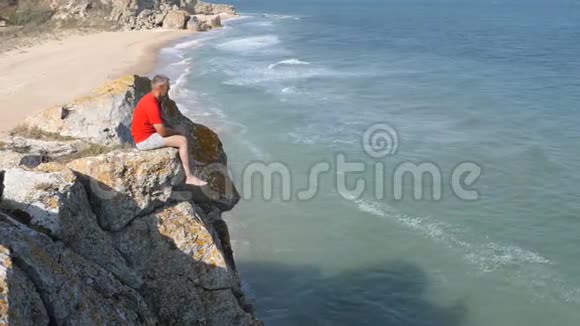人坐在悬崖边上波浪冲刷沙质海岸山脊在岩石上破碎了视频的预览图