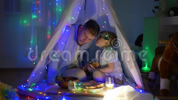 快乐的童年爸爸和儿子一起吃包子晚上坐在装饰着花环的帐篷里视频的预览图