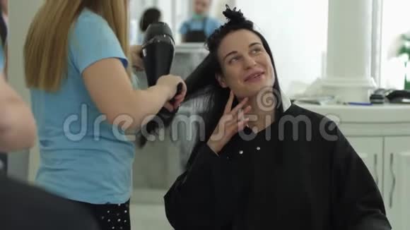 在美容院提供良好的服务快乐的女孩在用吹风机吹头时与理发师交谈视频的预览图