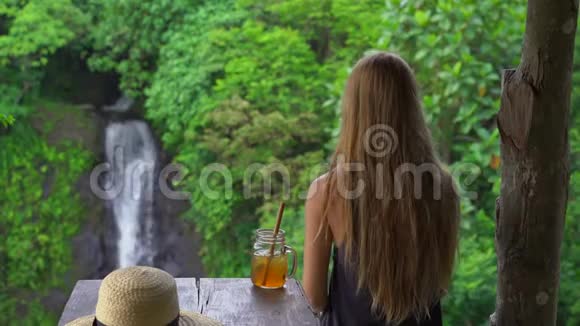 一位年轻的女士在一家美丽的咖啡馆里在峡谷和瀑布上有着史诗般的景色喝着冰茶前往巴厘岛视频的预览图