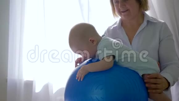 治疗师训练一个婴儿在房间里的蓝色充气大球背景上轻窗关闭视频的预览图