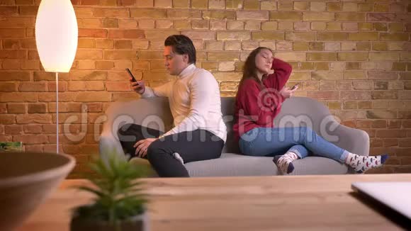年轻的白种人朋友坐在沙发上在家里的气氛中使用智能手机的肖像视频的预览图