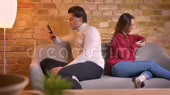 年轻的白种人夫妇背靠背坐在沙发上在家庭氛围中使用智能手机的肖像视频的预览图