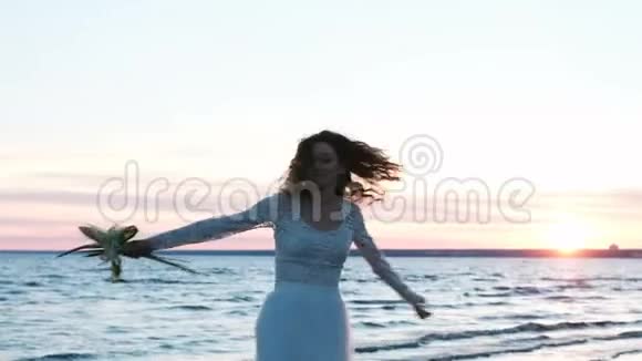 美丽的欧洲新娘带着一束手工制作的原始花束在日落时分在海上旋转在海滩上跳舞视频的预览图