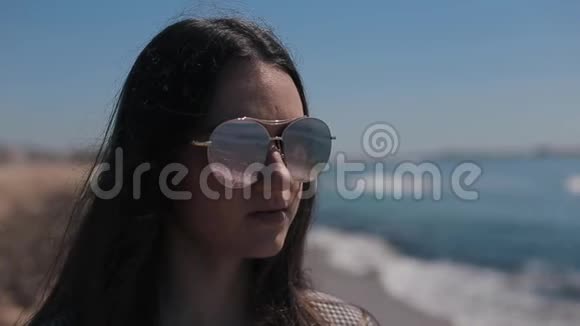 一幅美丽的女孩戴着太阳镜站在海边和海滩上的肖像模糊了背景大海的倒影视频的预览图