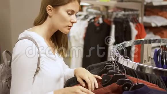 年轻女子在商场购物站在货架附近衣服选择夹克把蓝色外套捡起来视频的预览图