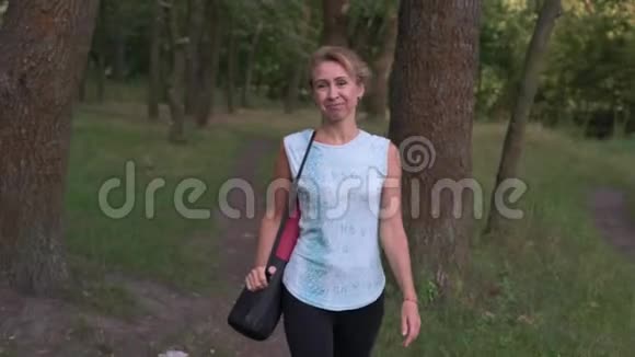 成熟的女人在森林中锻炼后继续前进用手在肩膀上握着瑜伽垫的空气亲吻来拯救我们视频的预览图