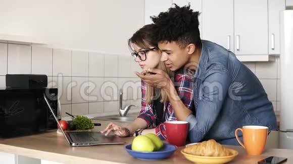 白人女孩和她20多岁的混血男朋友都在家里的厨房里用笔记本电脑上网视频的预览图