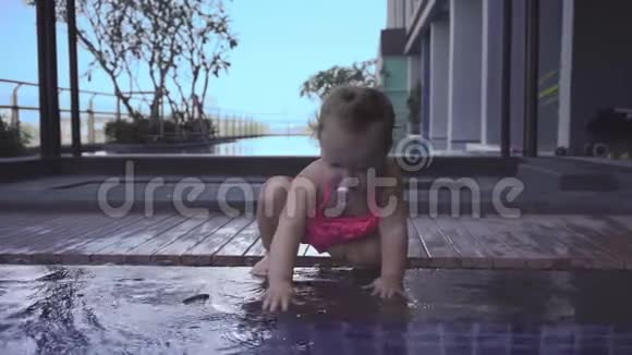 穿着粉红色泳衣的女孩坐在游泳池的浅滩上孩子从水池里扔石头慢动作视频的预览图