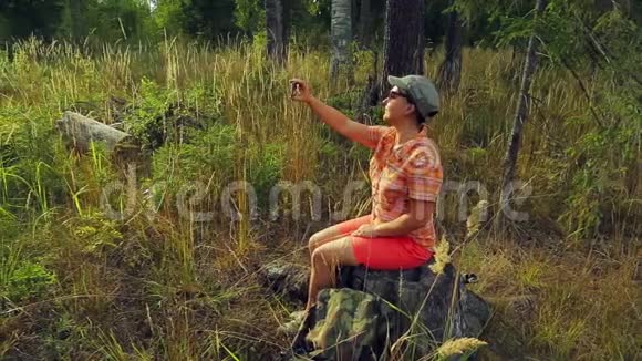 年轻的女游客戴着太阳镜和棒球帽坐在一棵倒下的树上在电话上拍照视频的预览图