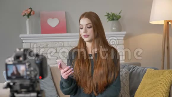 年轻的红发女孩博客写手微笑着对着镜头说话用智能手机阅读评论在家里安慰视频的预览图