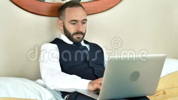 在酒店房间里躺在床上的男性商人在出差时使用笔记本电脑视频的预览图
