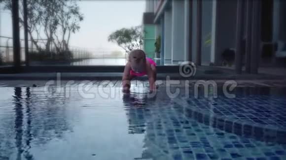 穿着粉红色泳衣的女孩坐在游泳池的浅滩上孩子从水池里扔石头慢动作视频的预览图