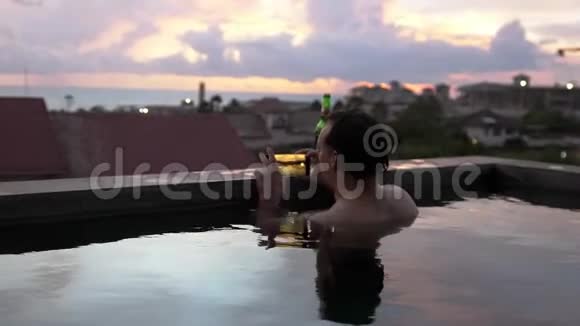 年轻人用智能手机在游泳池休息和喝啤酒时拍摄美丽的日落慢动作视频的预览图