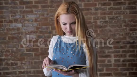 年轻迷人的金发女孩正在看书看着镜头微笑着砖色背景视频的预览图