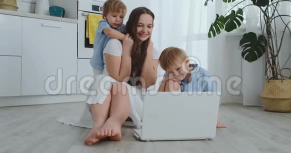 现代公寓爱妈妈和两个小儿子坐在客厅的地板上看着笔记本电脑屏幕视频的预览图