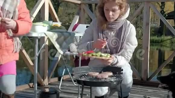 一个穿着暖和衣服的年轻女子在烤蔬菜遛狗在河边的木桥上野餐视频的预览图