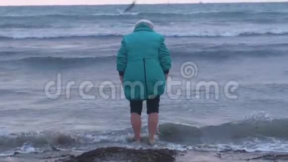 穿着帽子和夹克的老妇人在日落时正在海水中散步后景视频的预览图