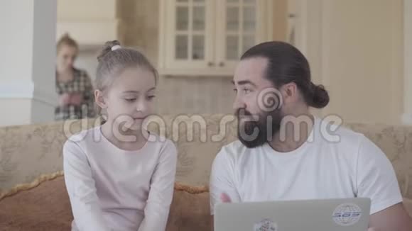 爸爸带着可爱的女儿和笔记本电脑坐在客厅的沙发上谈论着一些有趣的事情视频的预览图