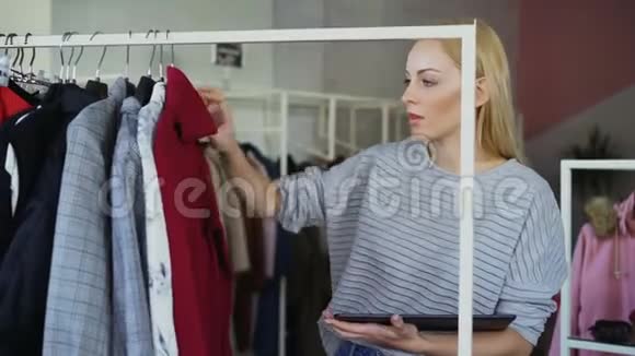 年轻的女商人正在检查铁轨上的服装并在她的服装店里用平板电脑工作她正在检查价格视频的预览图