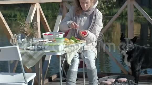 年轻女子穿着温暖的衣服在河岸上的木桥上野餐切蔬菜一只狗在附近玩耍视频的预览图