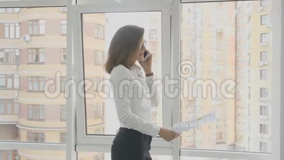 办公室工作人员戴眼镜的商务女士站在办公室里看文件和打电话视频的预览图
