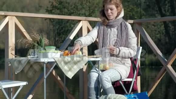 年轻的女人穿着暖和的衣服在河岸上的木桥上野餐在桌子上放水果周末寒冷视频的预览图