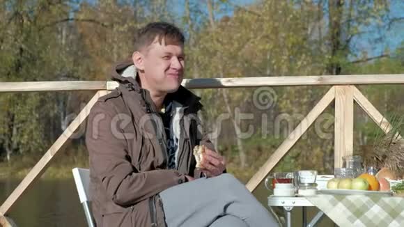 成熟的男人穿着温暖的衣服坐在桌子旁吃着汉堡包微笑着从热水瓶里倒茶野餐视频的预览图
