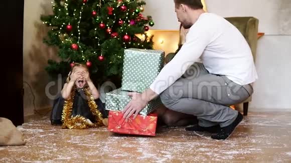 可爱的小女孩坐在圣诞树下闭着眼睛而她的父亲正带着礼盒视频的预览图