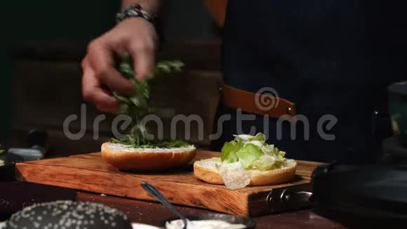 一位身穿蓝色围裙的男厨师的特写从汉堡包中取出多余的绿色库存录像筹备视频的预览图