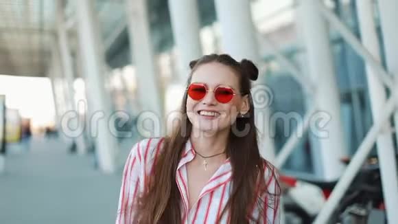 戴着红色太阳镜的快乐时尚模特自信地沿着购物中心附近的街道走着夏季视频的预览图