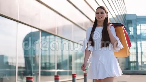 漂亮的时装模特穿着白色连衣裙在现代玻璃建筑前摆着购物袋慢动作视频的预览图
