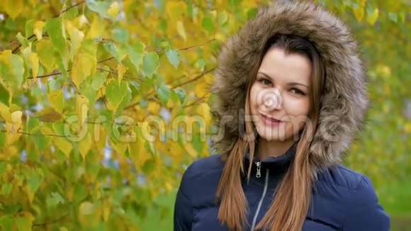 女孩在秋天的城市公园散步穿夹克衫的年轻女子在秋树背景上戴着毛皮帽女孩走在里面视频的预览图