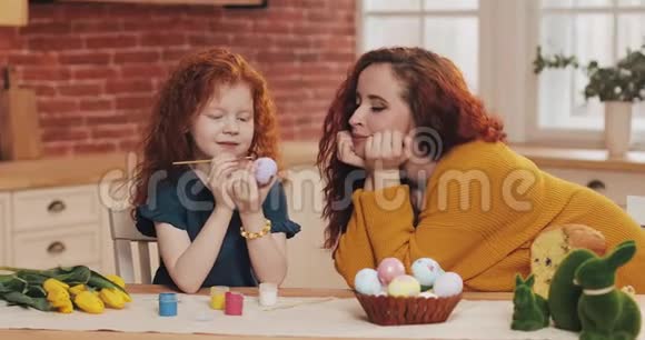 妈妈和她的小女儿正在准备复活节小女孩在舒适的厨房里画复活节彩蛋微笑的母亲视频的预览图