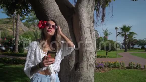 一个漂亮的女人站在棕榈树附近手里拿着电话视频的预览图