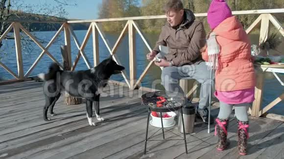 父亲和女儿成熟的男人和女孩穿着温暖的衣服坐在桌子旁喂狗在河岸野餐视频的预览图
