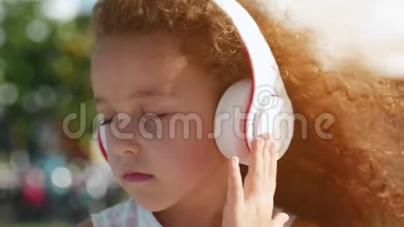 可爱的白种人卷发小女孩穿着白色连衣裙坐在公园里戴着白色耳机听音乐看着视频的预览图