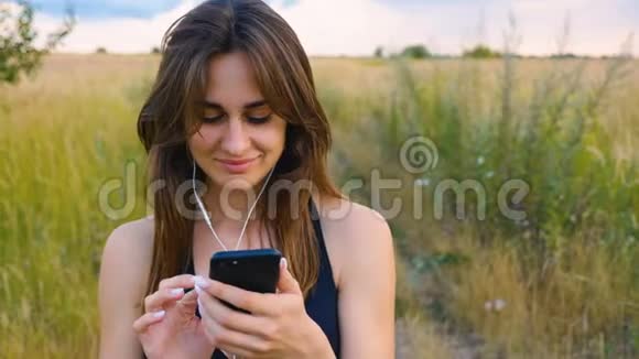 快乐的年轻女性使用智能手机和耳机在日落时站在田野健康的生活方式女运动员使用视频的预览图