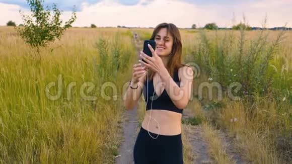 快乐的年轻女性使用智能手机和耳机在日落时站在田野健康的生活方式女运动员使用视频的预览图