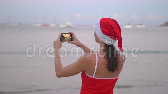 日出时身穿圣诞帽和红色泳衣的女士在海滨用智能手机手机拍照视频的预览图