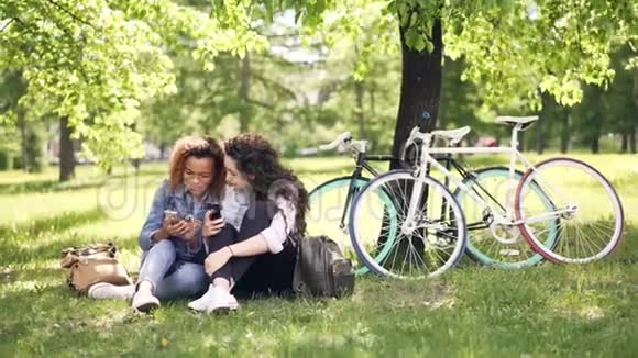 开朗的年轻女性白人和非裔美国人在公园里聊天和使用智能手机女孩在看屏幕视频的预览图