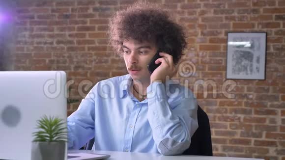 年轻的书呆子商人留着卷发和胡子在电话上交谈在笔记本电脑上工作坐着现代化的办公室视频的预览图