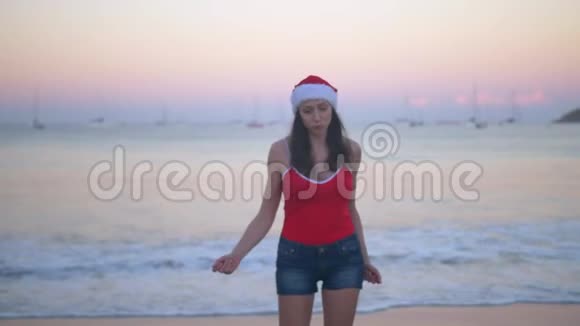 在热带天堂海滩日落时穿着圣诞帽和红色泳衣的无忧无虑的女人跳舞和跳跃快乐视频的预览图