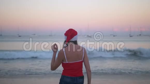 在热带天堂海滩日落时穿着圣诞帽和红色泳衣的无忧无虑的女人跳舞和跳跃快乐视频的预览图