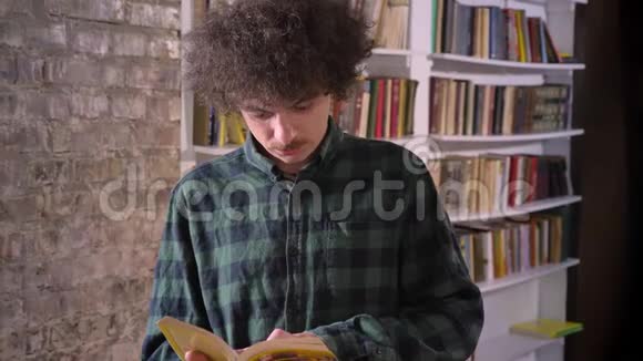 聪明的书呆子学生卷发在图书馆看书站着书架在后台视频的预览图