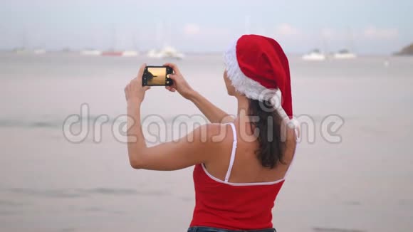 日出时身穿圣诞帽和红色泳衣的女士在海滨用智能手机手机拍照视频的预览图