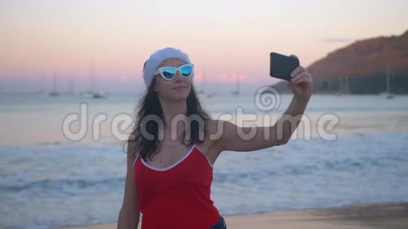穿着红色圣诞帽的有趣女人在热带海滩度假时拍照视频的预览图
